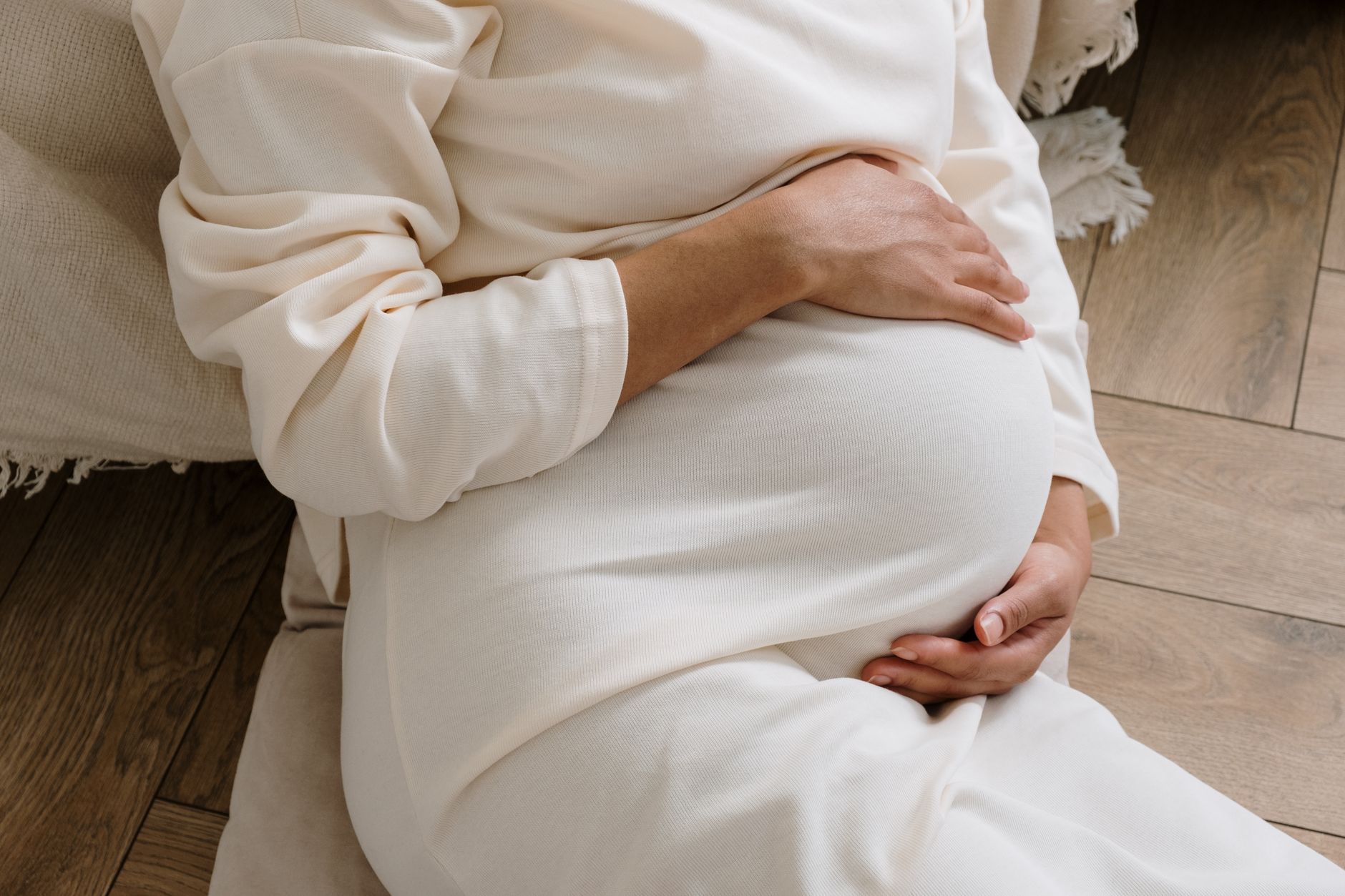 O femeie însărcinată într-o rochie albă tricotată, stând pe podea, cu mâinile pe burtă; fața nu se vede